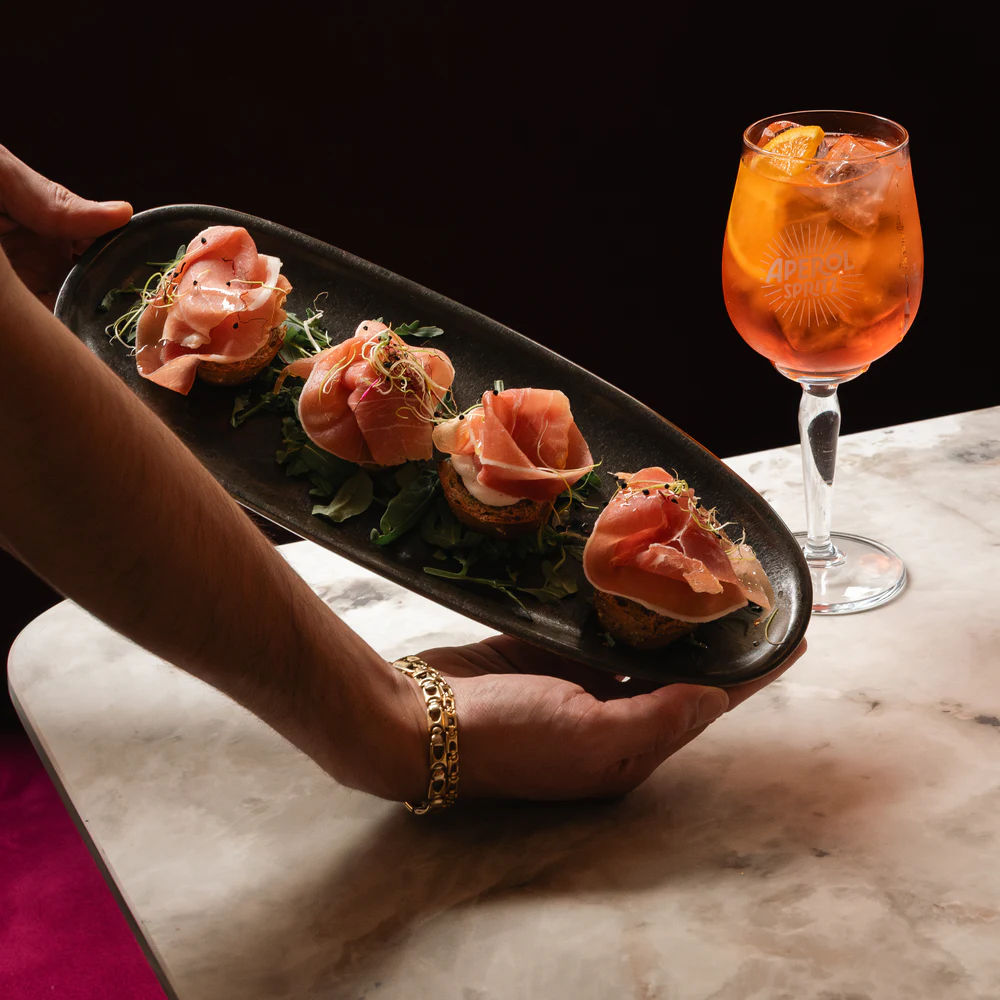 Ein Brett mit Crostini wird an einem Aperol Spritz auf einer Marmortheke vorbeigetragen.
