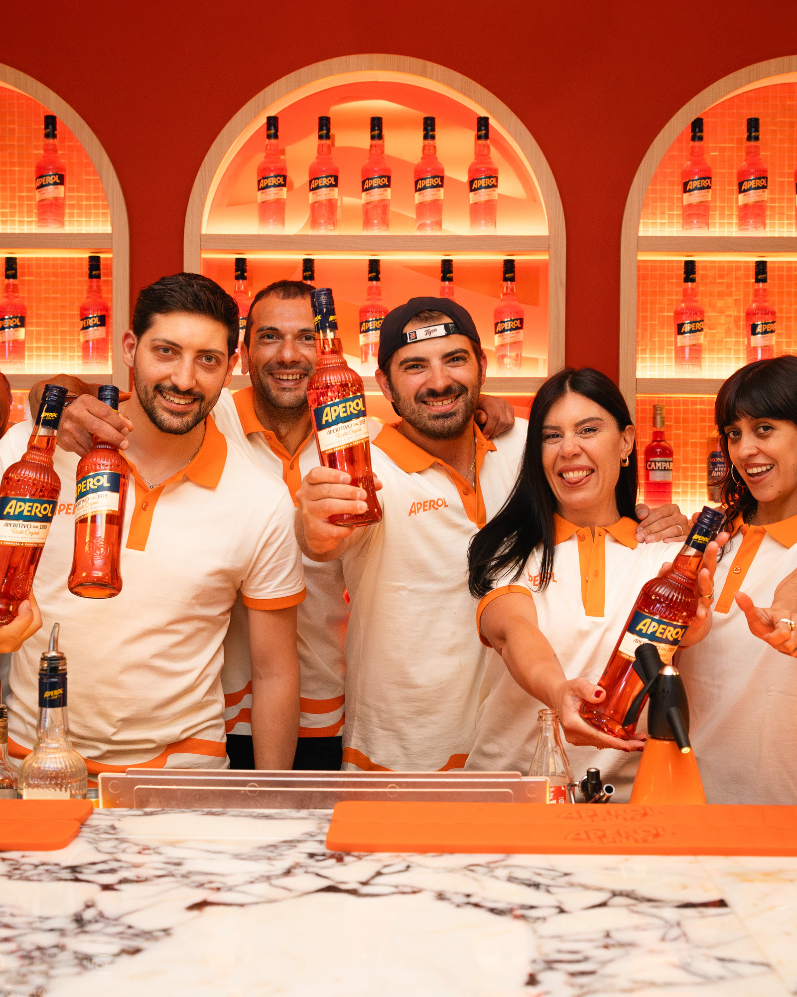 Das Aperol-Team hinter der Bar, die Flaschen erhoben, unter der beleuchteten Flaschenwand