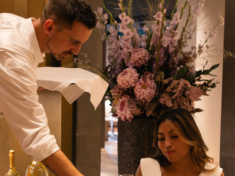 Ein Kellner bedient einen sitzenden Gast an einem Tisch mit Blumen und Champagner